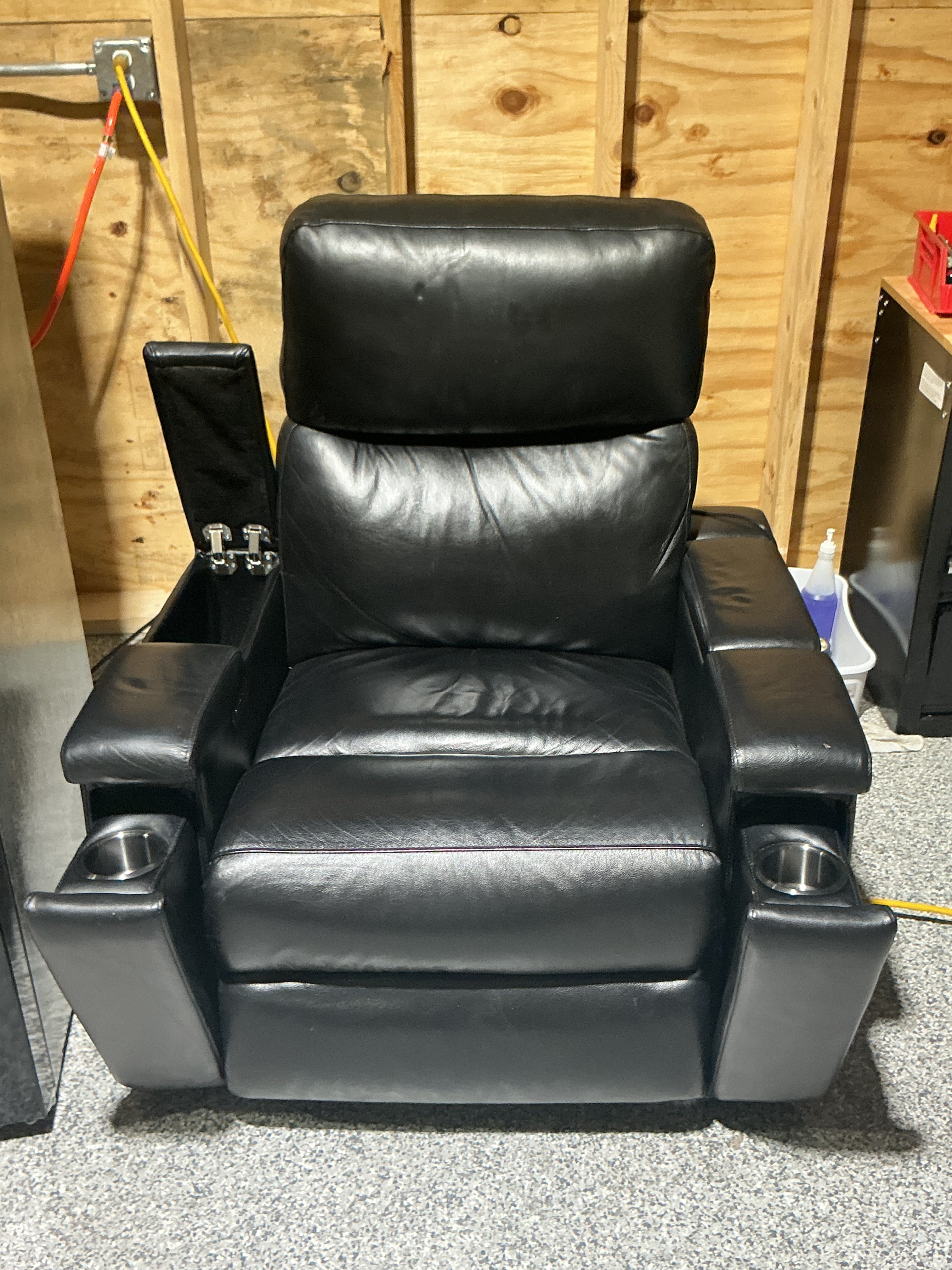 Black leather recliner chair with armrests and cup holders, set against a wooden wall.